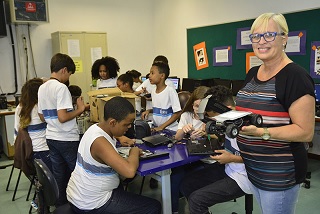 Em primeiro plano, a professora Maria Cristina Zamith exibe um roboô construído em sala de aula. Ao fundo, os estudantes trabalham com outras peças.