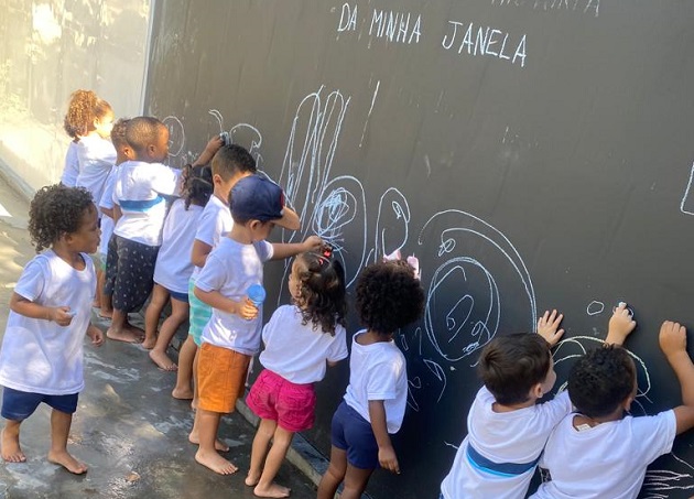 Foto. Crianças pequenas desenham com giz em uma parede negra. No alto da parede está escrito 