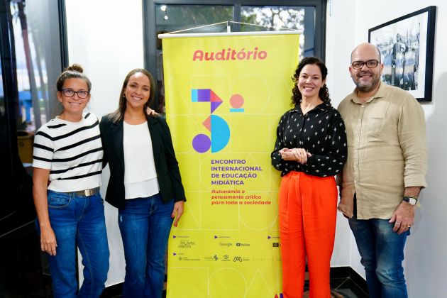 Foto. Quatro representantes da MultiRio estão em pé, lado a lado, posando e sorrindo para a foto. Entre eles o banner vertical do evento.