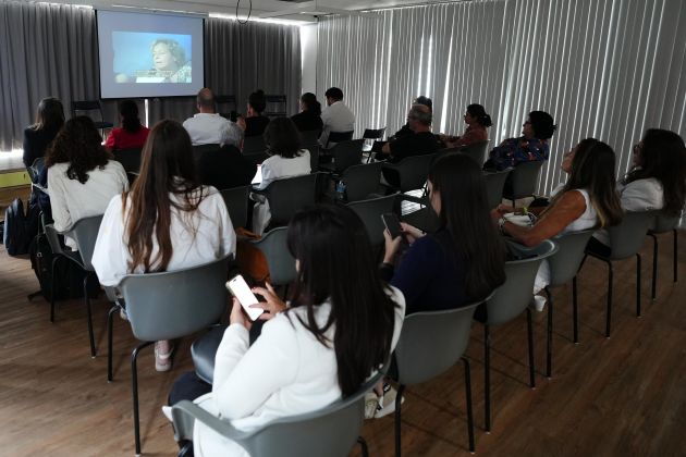 Foto. Pessoas estão sentadas em cadeiras em uma sala escura com um telão.