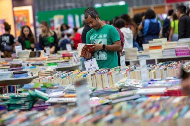 Foto. Espaço com inúmeros livros expostos em mesas, cobrindo uma grande área, o que sugere uma feira de livros. Ao fundo, de forma desfocada, várias pessoas de diferentes gêneros e idades estão envolvidas na atividade de escolher livros. Em primeiro plano, homem segura e folheia livro.