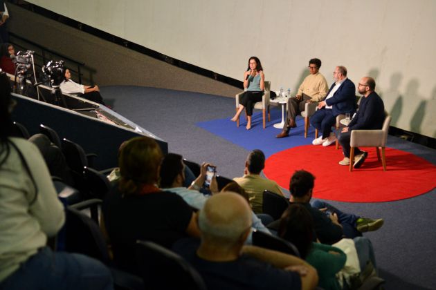Foto. Em primeiro plano desfocado, diversas pessoas de costas sentadas em uma plateia. Ao fundo, com foco, quatro pessoas sentadas em cadeiras em um palco.